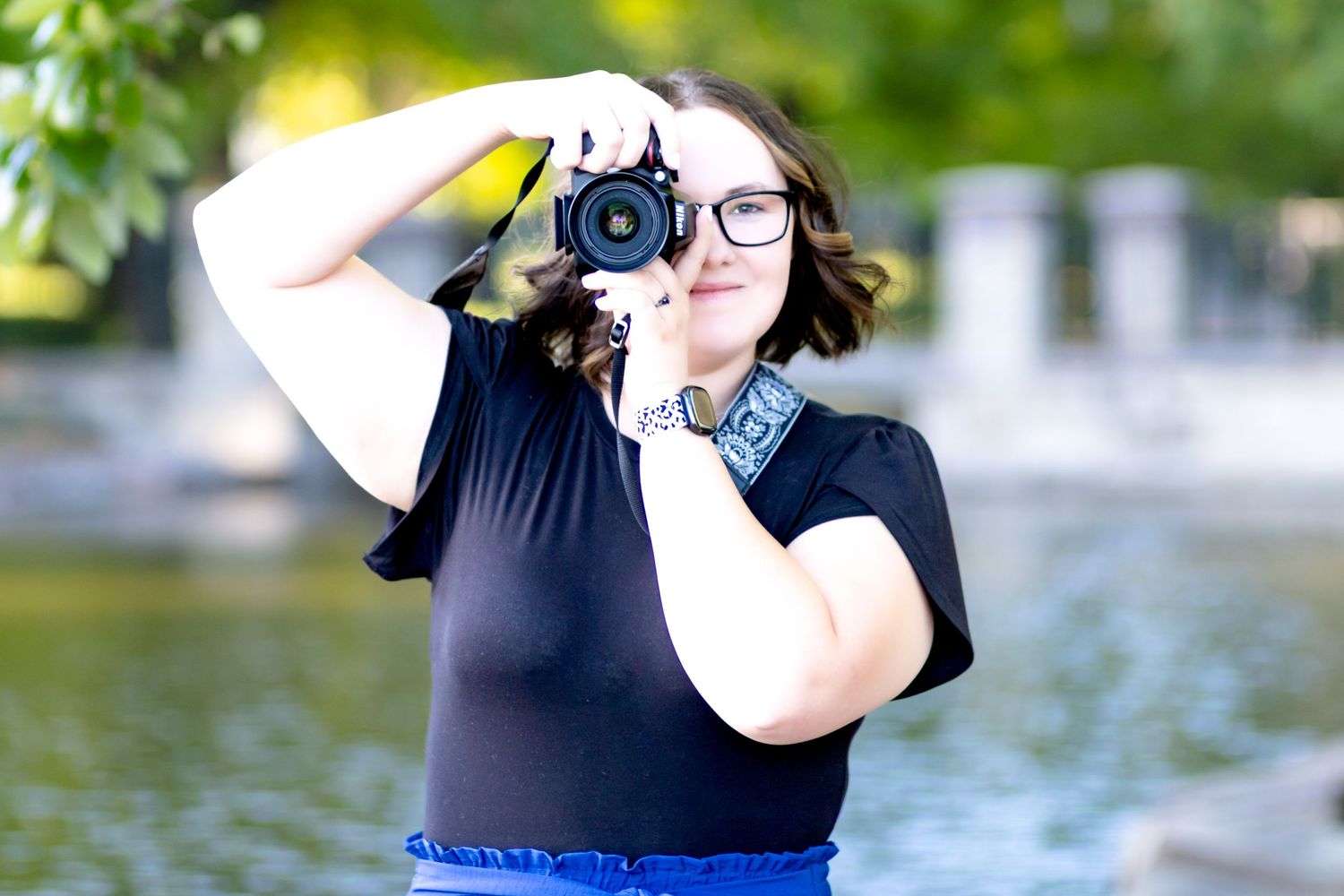 A photographer wearing glasses takes a photo with a DSLR camera near a lake on a sunny day.