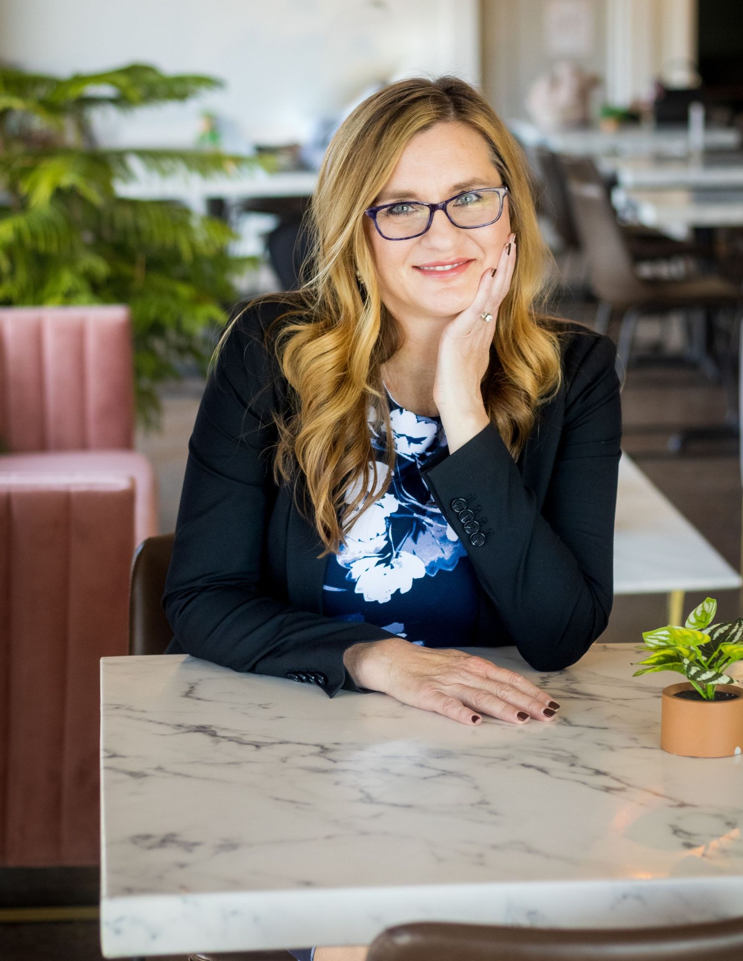 Professional woman in glasses and blazer sits at marble table with houseplants in modern office setting.