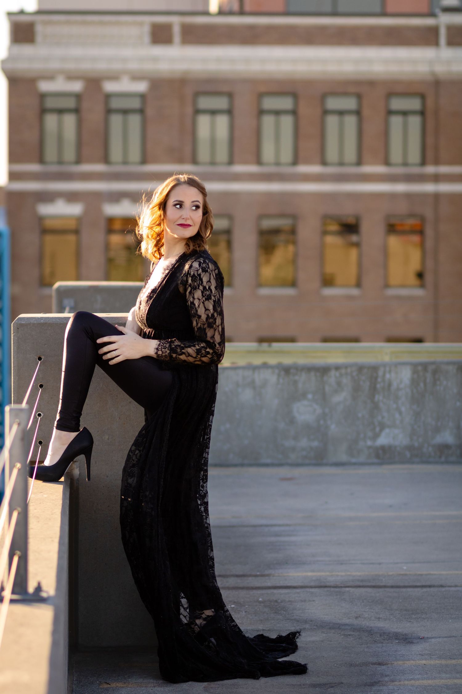 Professional urban rooftop portrait in black lace dress and heels with soft golden lighting and industrial backdrop.