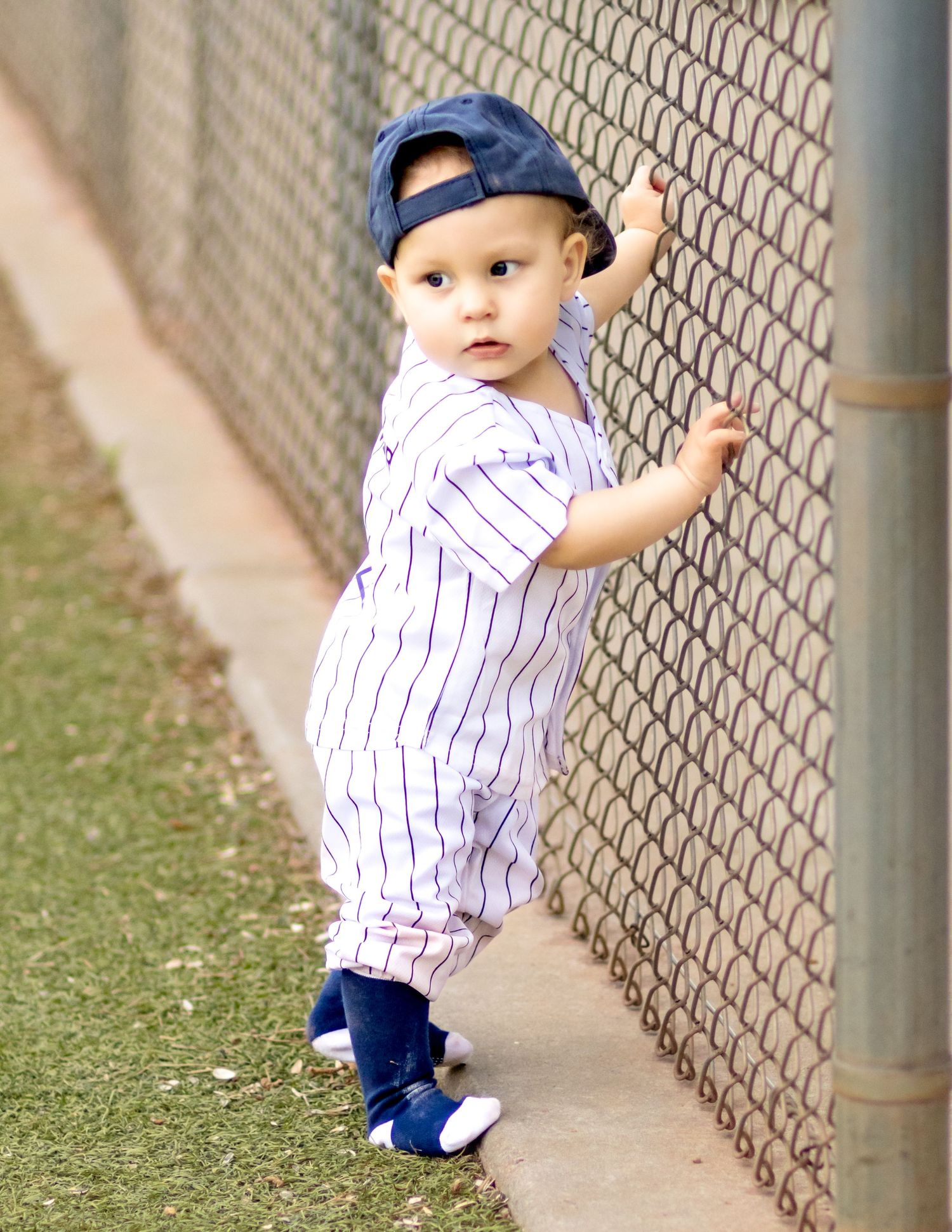 A young toddler wearing baseball attire and cap stands by a chain link fence on a baseball field.
