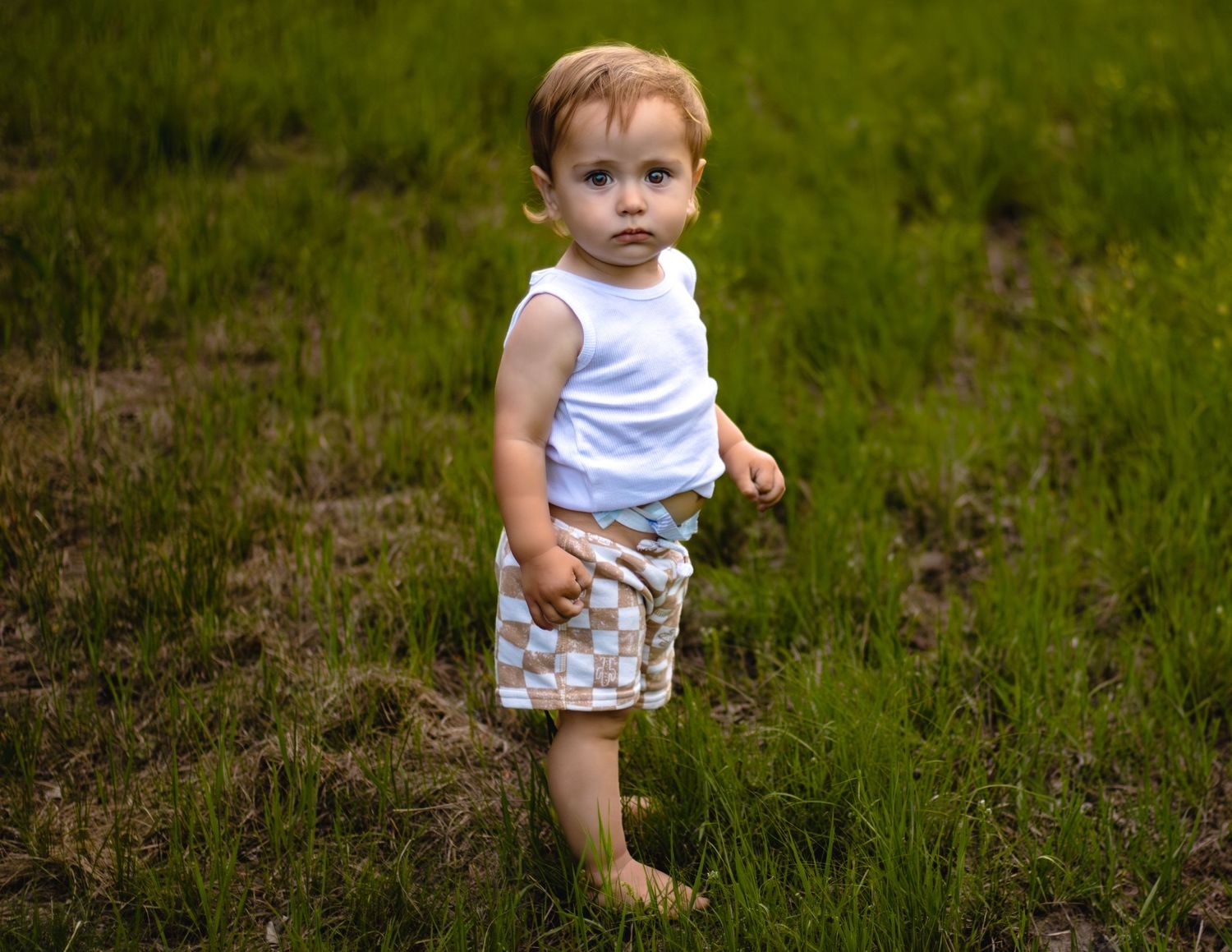 Small child wearing a white tank top and camouflage shorts stands in a grassy field on a summer day.