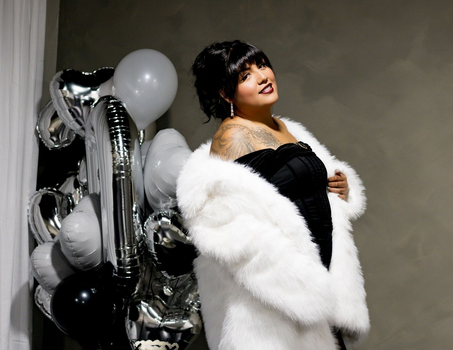 Glamorous figure in black dress and white fur stole poses elegantly beside silver and white party balloons.