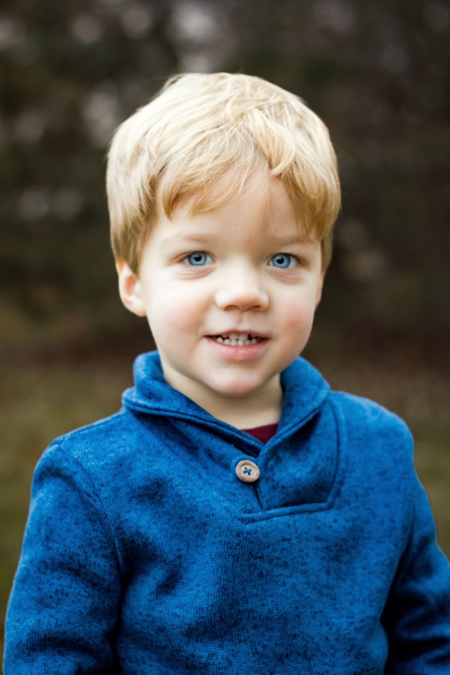 A young child wearing a blue button-up sweater smiles while posing for a portrait outdoors.