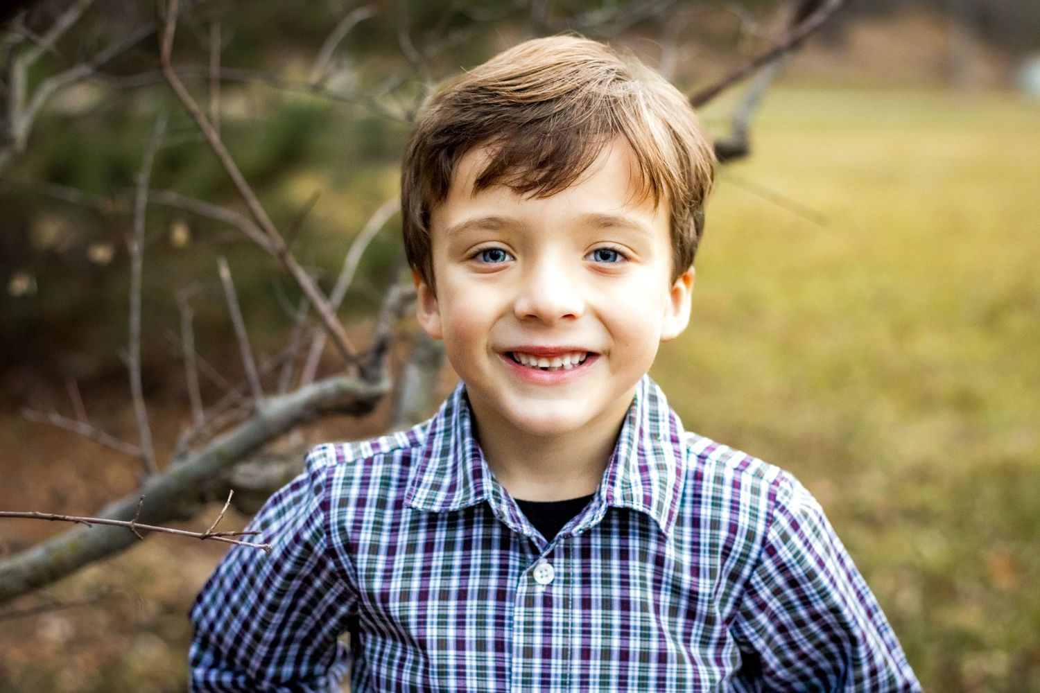 A young child in a blue plaid button down shirt smiles brightly outdoors in nature.