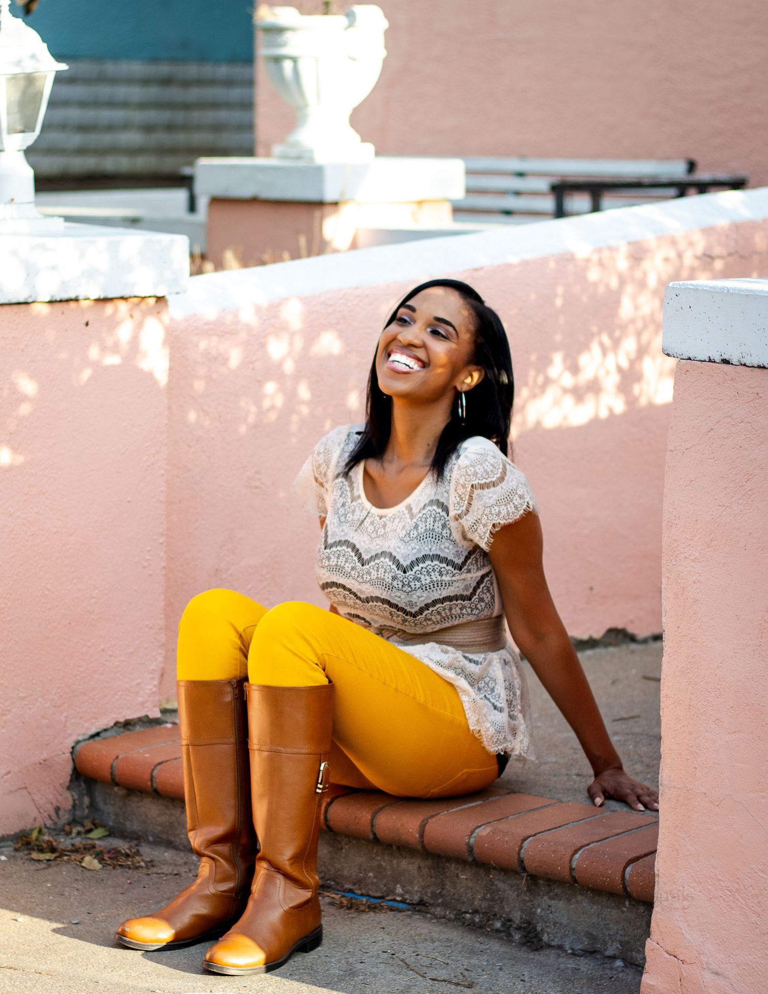 A person wearing a lace top, yellow pants and brown boots sits smiling on a pink wall in natural sunlight.