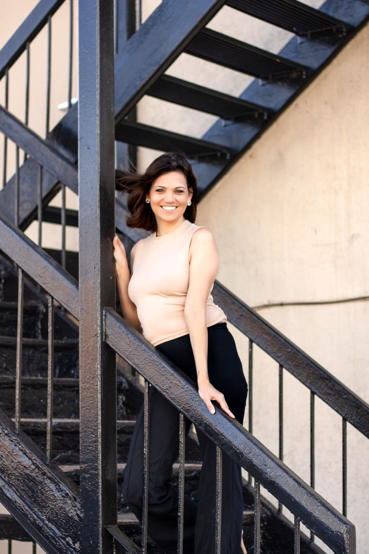 A woman in a beige crop top stands poised on black metal stairs against a light colored wall with natural lighting.