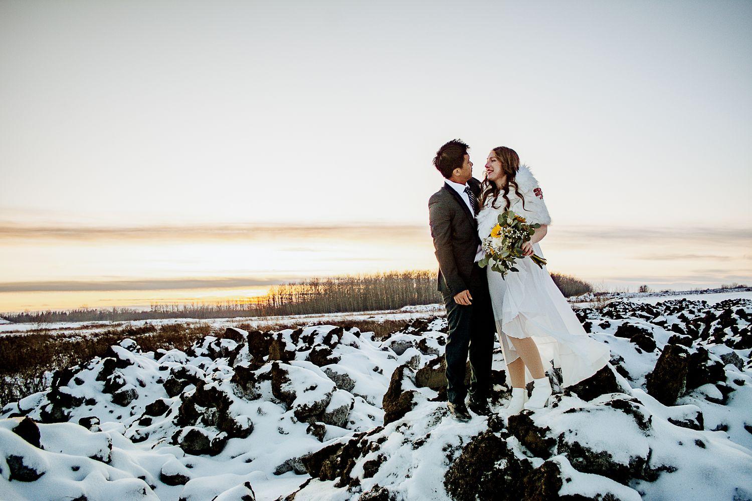 Winter wedding couple standing together on snowy rocky terrain at sunset.
