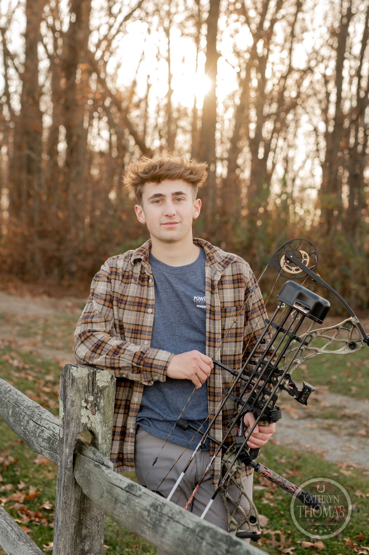 An archer poses with their compound bow against a wooden fence at sunset in autumn.