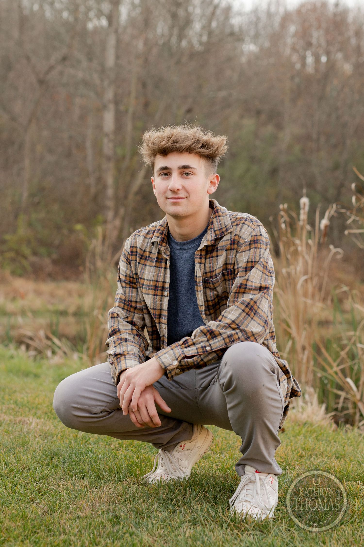 A person in casual attire crouches by a lake surrounded by autumn foliage and cattails.