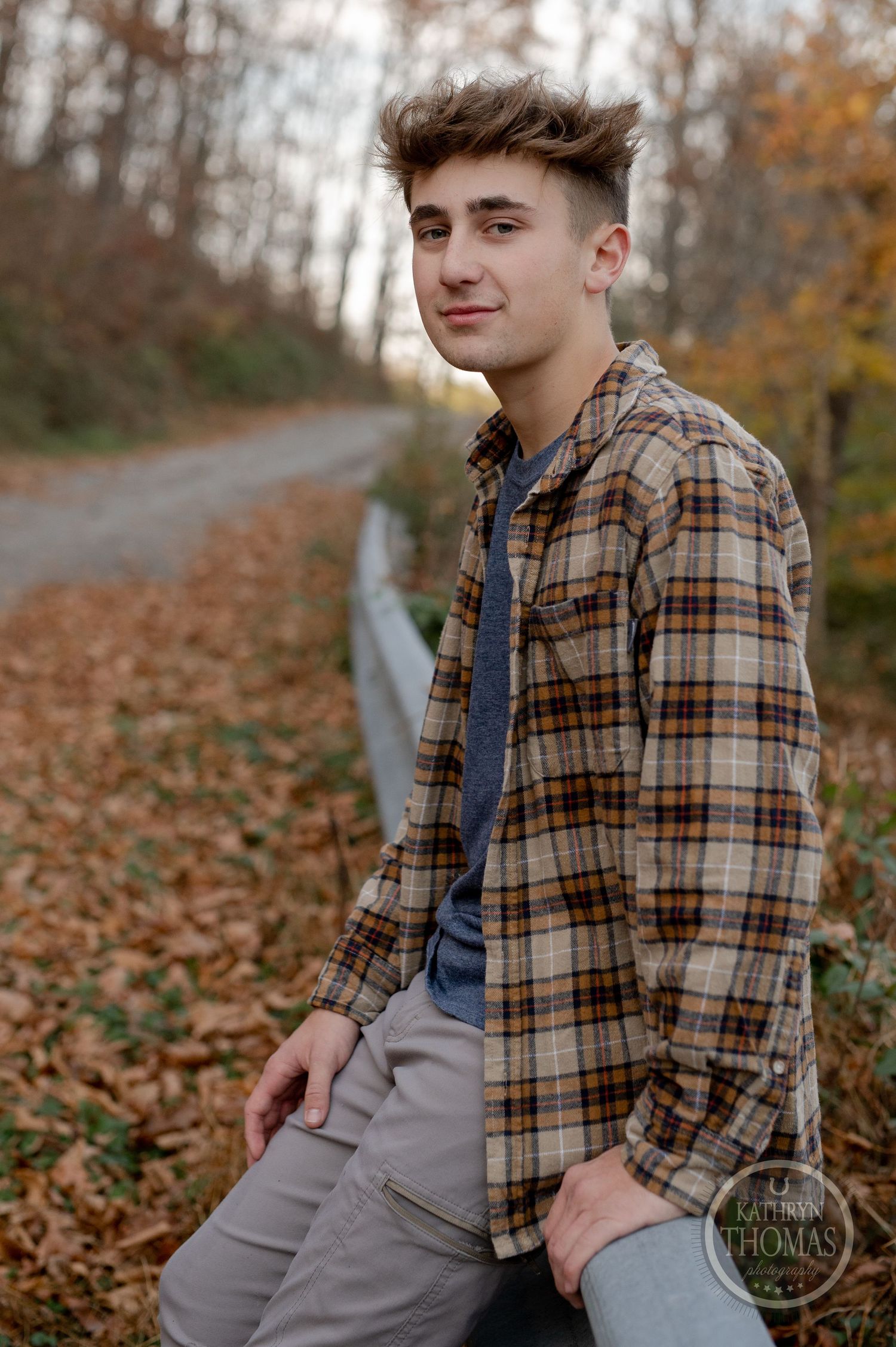 A person in a brown plaid shirt poses casually against a guardrail surrounded by fall foliage.