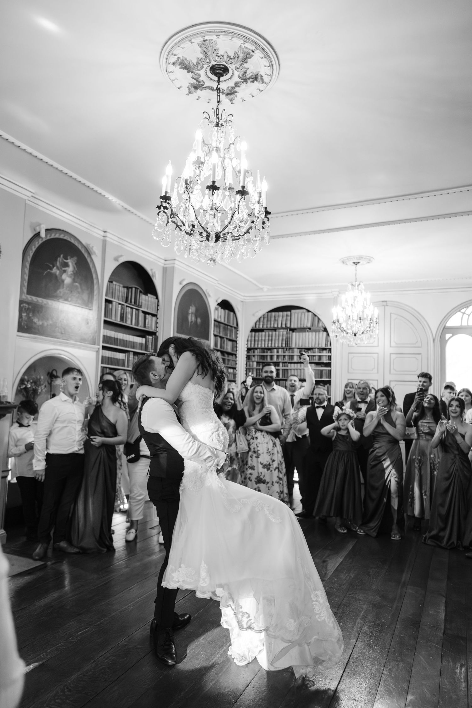 Black and white photo of bride and groom sharing first dance kiss, guests applauding in elegant ballroom with chandeliers.