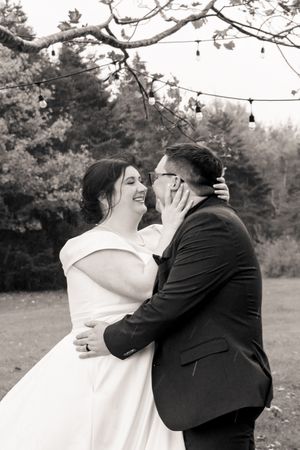 A black and white photo of a romantic moment between a couple in wedding attire beneath tree branches.