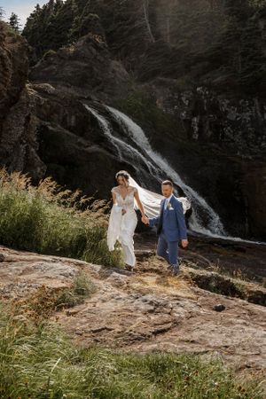 Two people walk together on rocky terrain near a dramatic cascading waterfall in a forested mountain landscape.