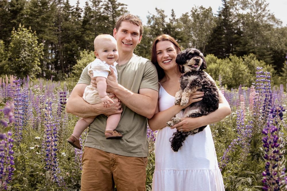 A couple stands with their baby and dog in a field of purple lupine flowers on a summer day.