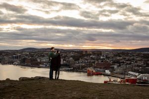 Two silhouettes watch the sunset over a scenic harbor town with boats and buildings nestled beneath cloudy skies.