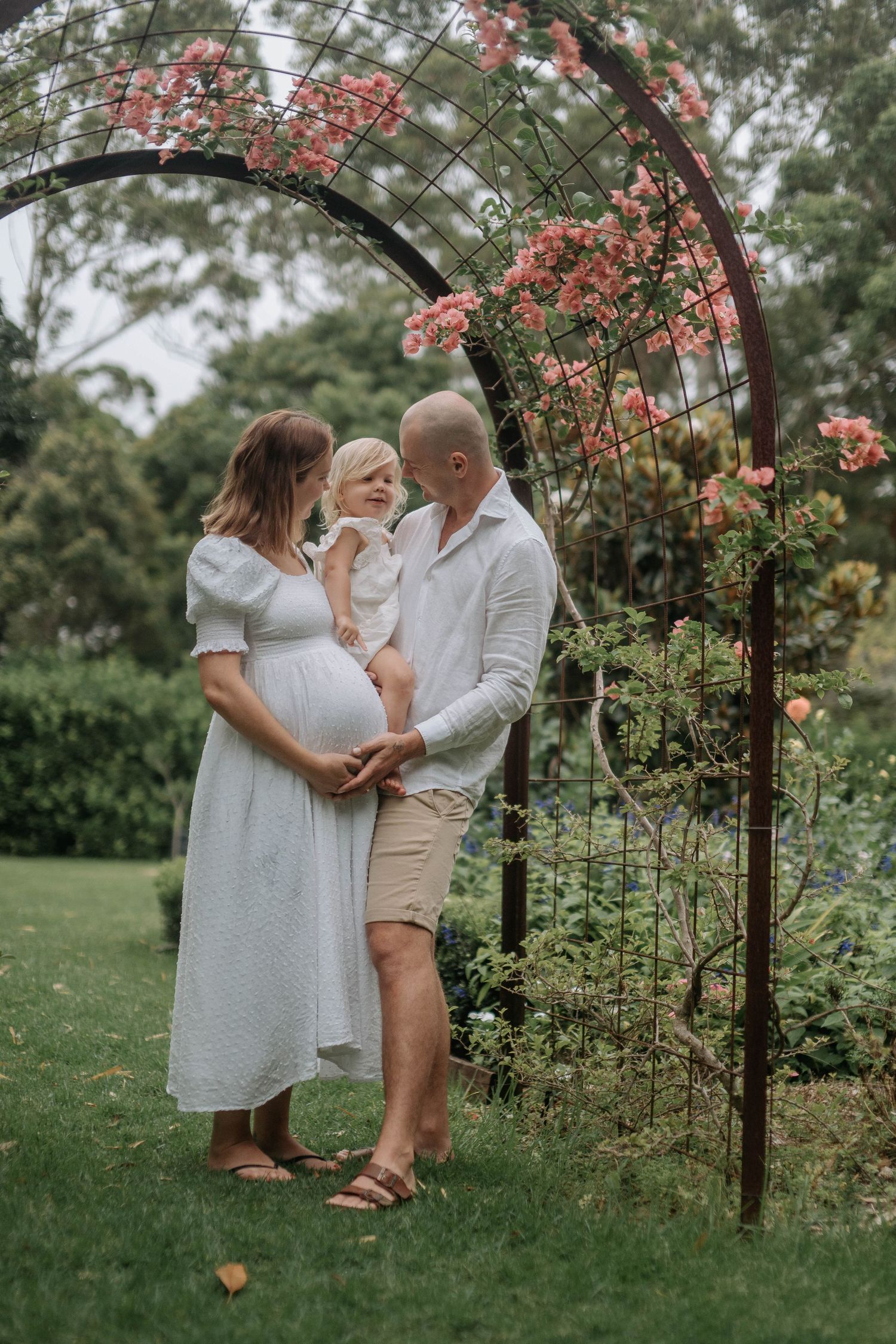 Expectant couple in white sharing intimate moment under soft sky.