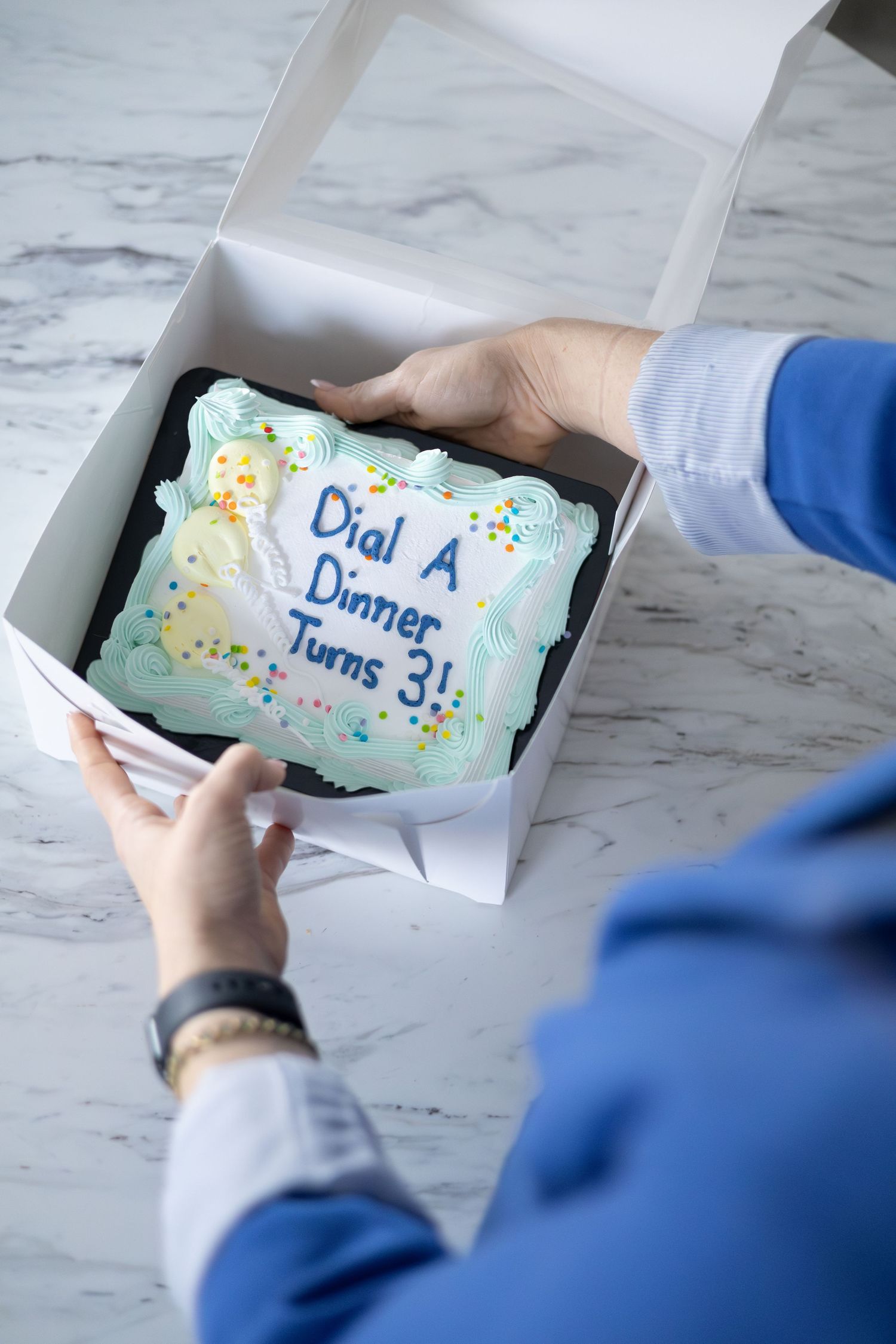 Hands opening a white bakery box containing a decorated cake with text 'Dial at Dinner turns 3!' on marble countertop.