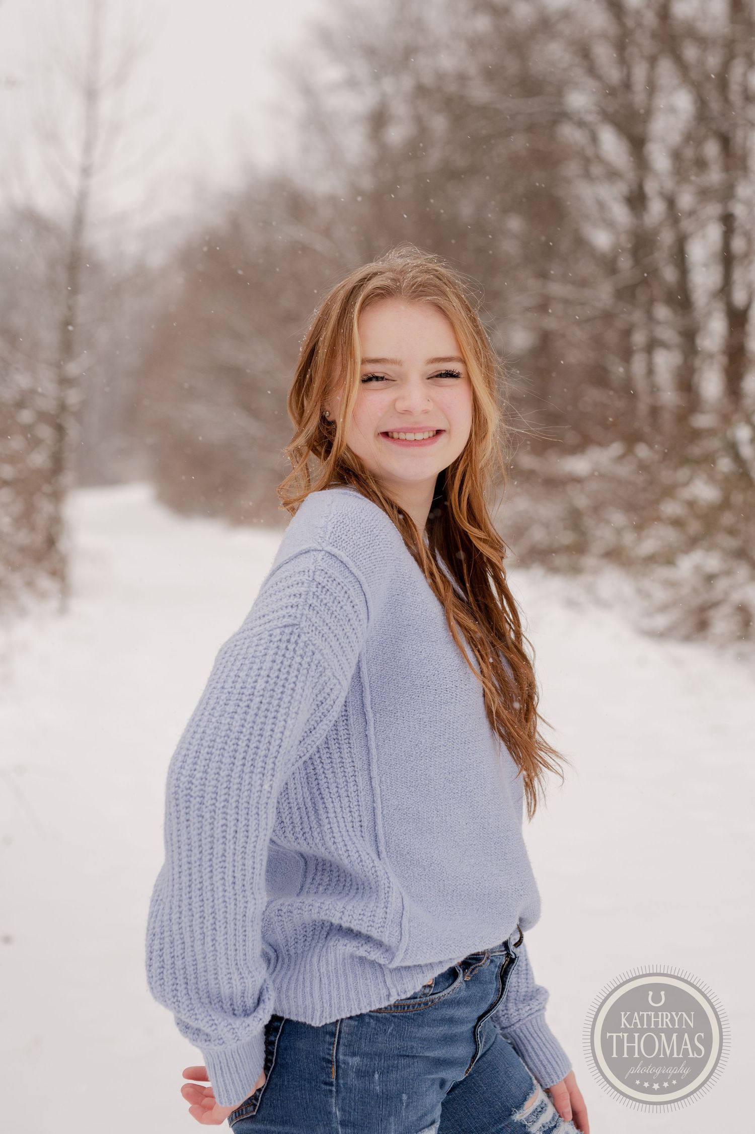 Someone wearing a cozy gray sweater smiles while posing on a snow-covered winter path.