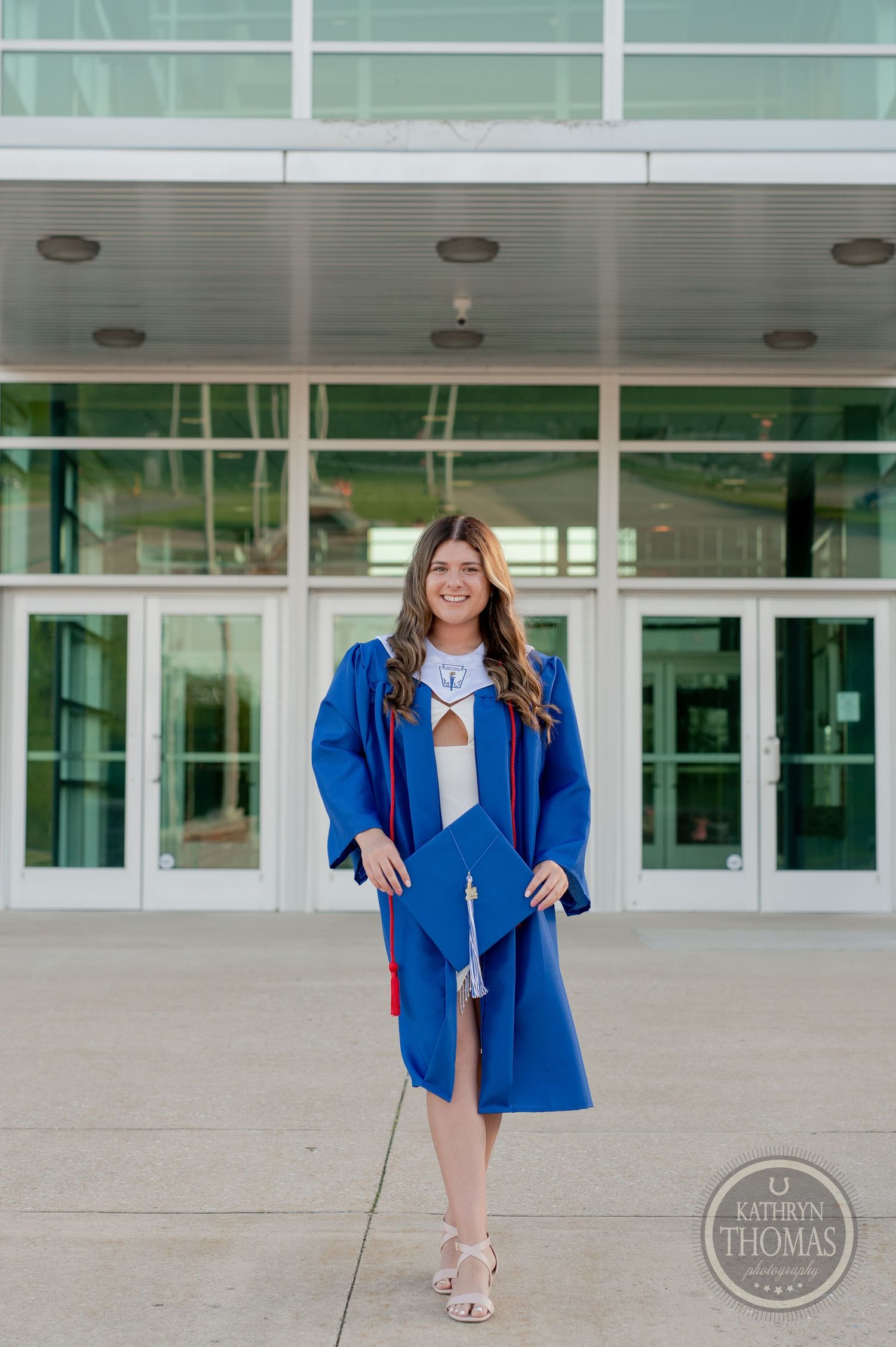 Graduate in blue cap and gown poses confidently in front of modern school entrance.
