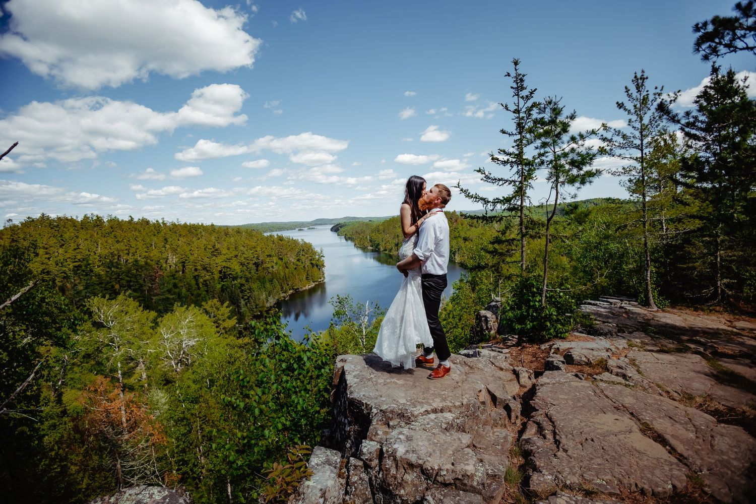 Newlyweds embrace on rocky cliff overlooking scenic forest lake landscape.