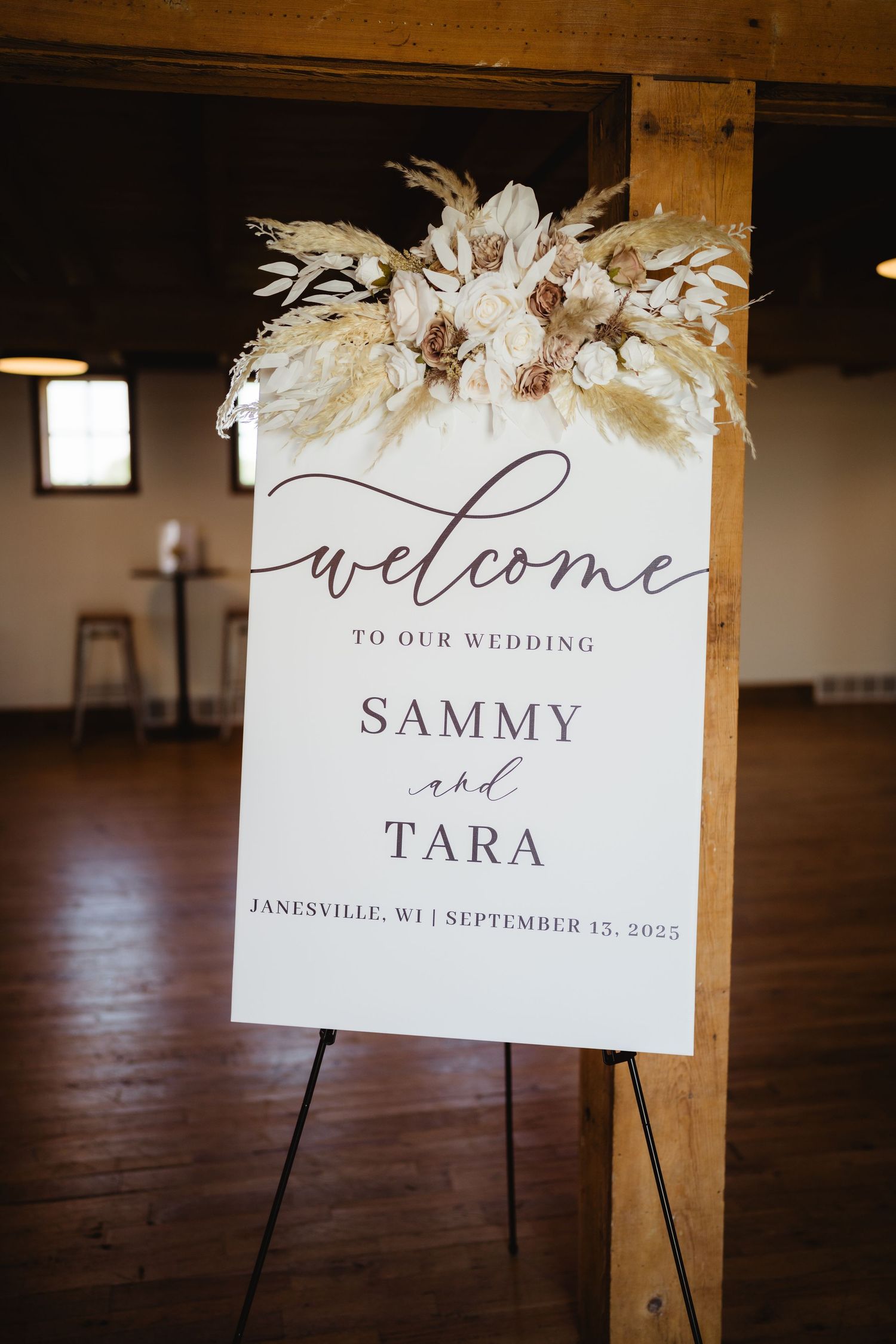 White wedding welcome signs with pampas grass decorations displayed on easels in a rustic venue.