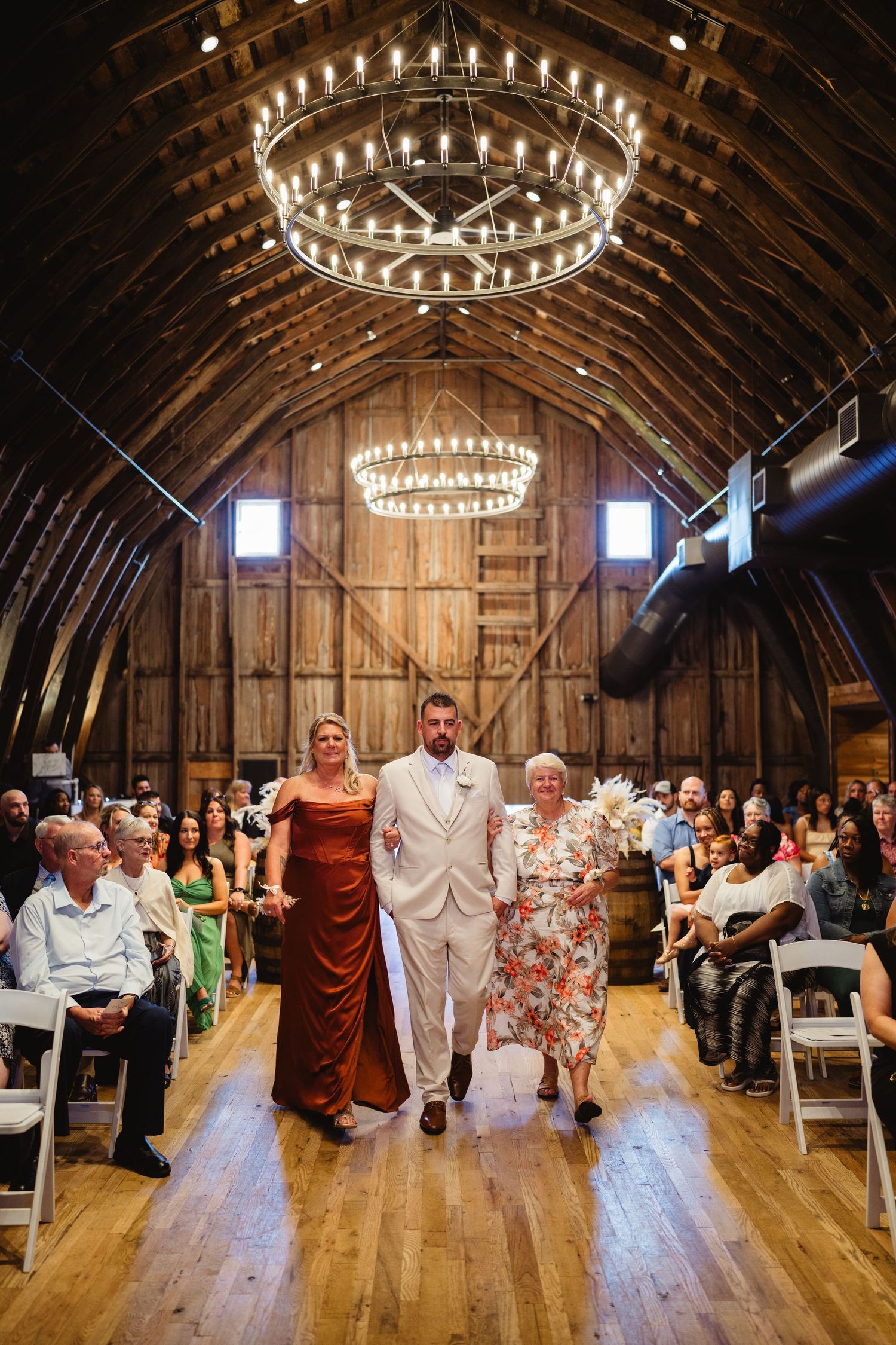 Wedding reception in a rustic barn with chandeliers and guests celebrating on a wooden dance floor.