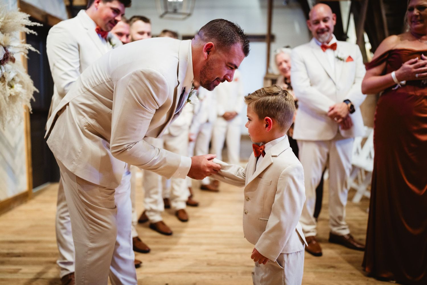 People in white suits share a tender moment with a young boy in a matching white outfit on a wooden dance floor at an event.