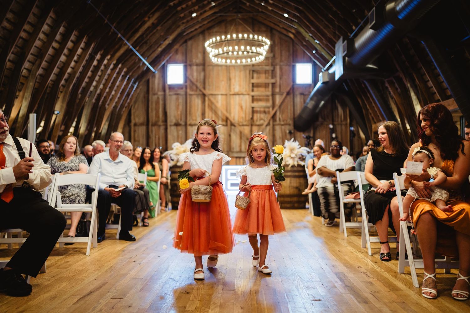 Two people in red skirts walk down wooden aisle at rustic barn wedding ceremony while guests watch from white chairs.