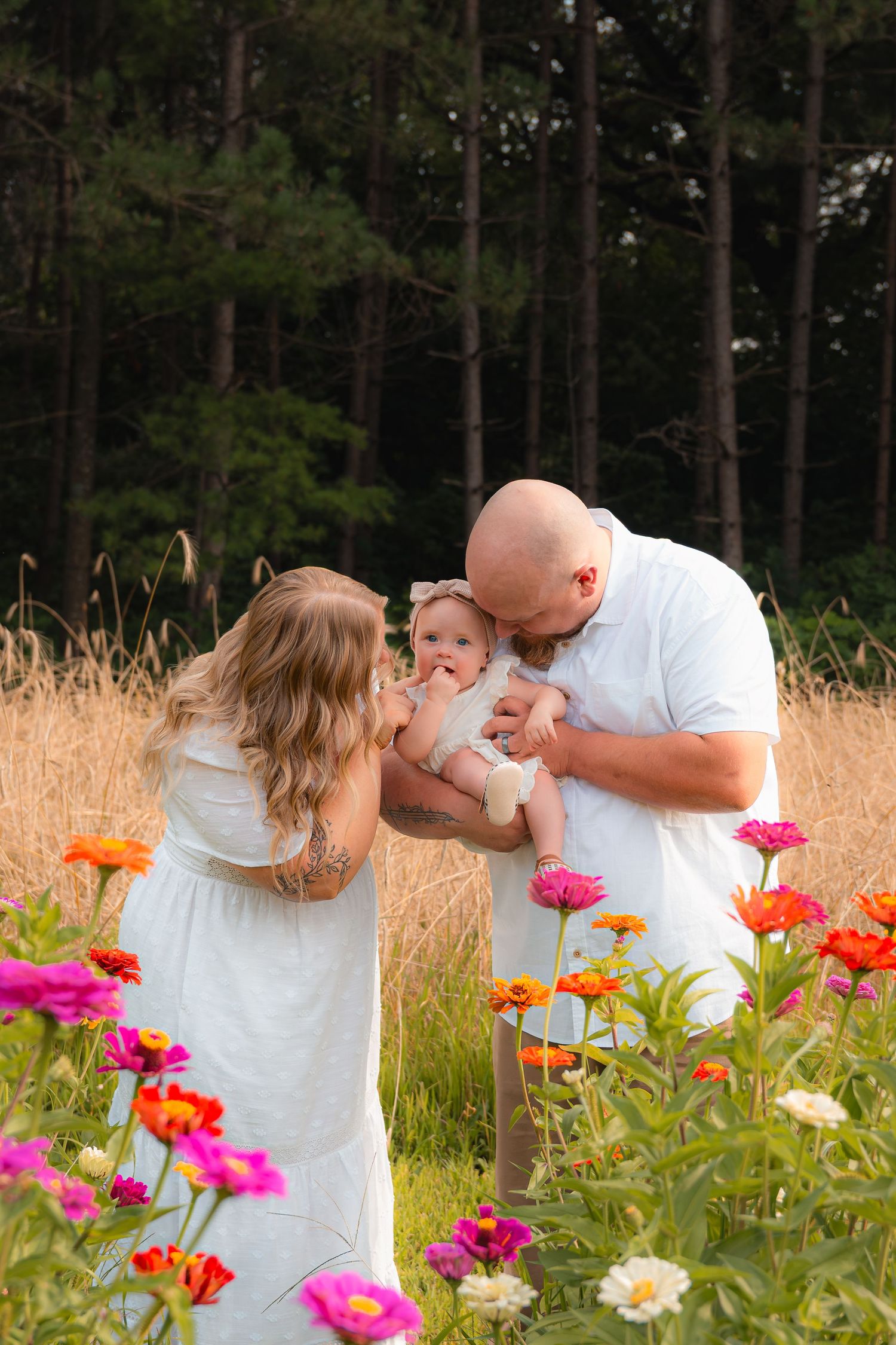 Family in white outfits kissing baby in colorful zinnia flower field with tall pine trees backdrop.