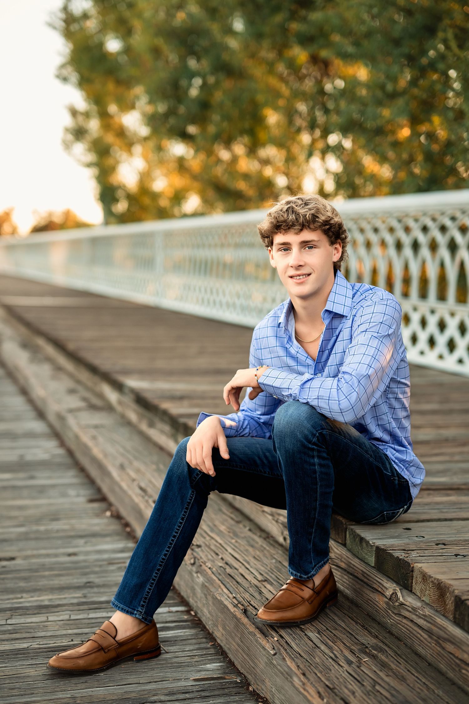 Senior boyposes editorially in a blue checkered shirt and jeans sitting on wooden boardwalk during golden hour.