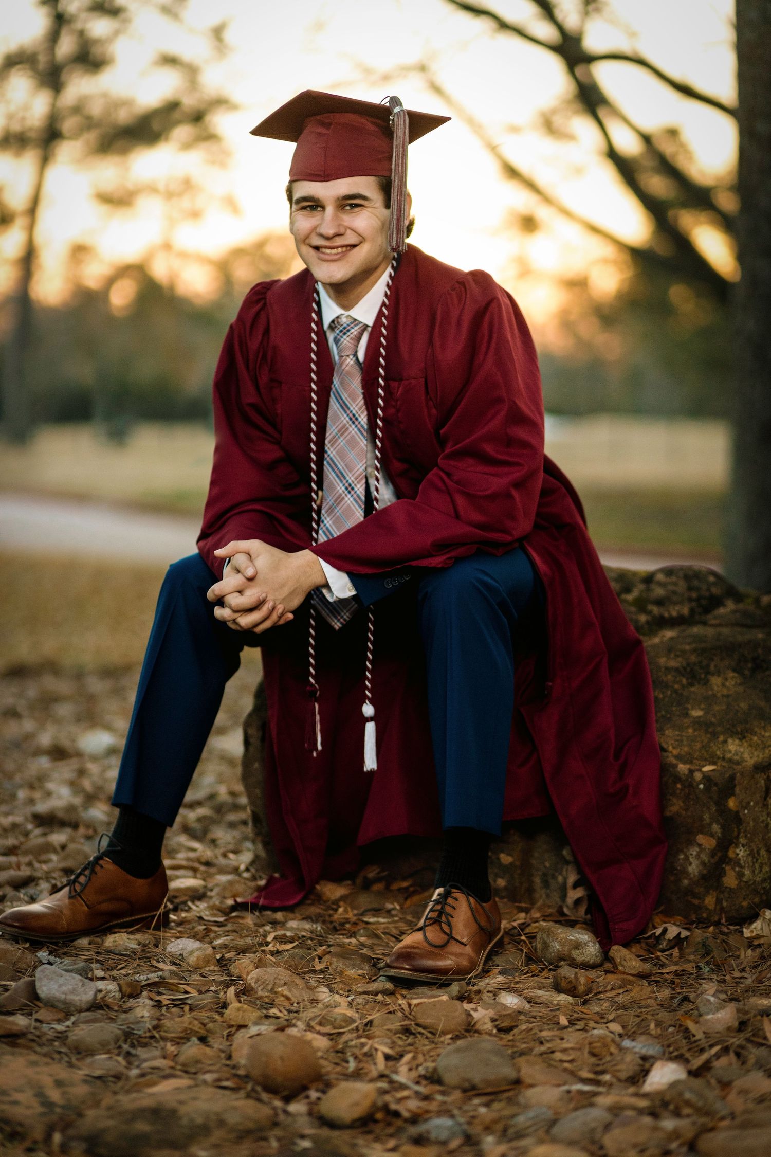 Graduate in maroon cap and gown sits on stone wall surrounded by autumn leaves at sunset.