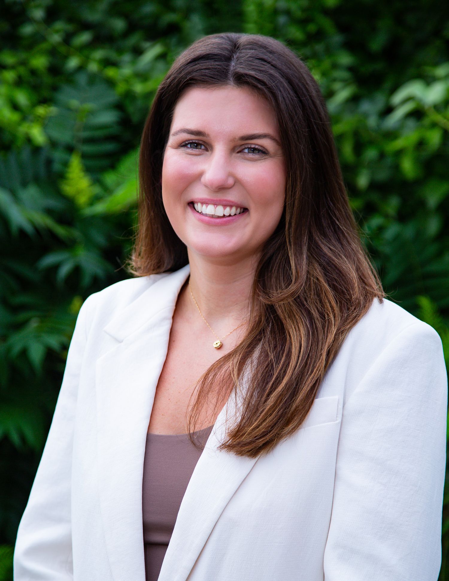 Professional headshot portrait with a bright smile wearing a white blazer against a green leafy background outdoors.