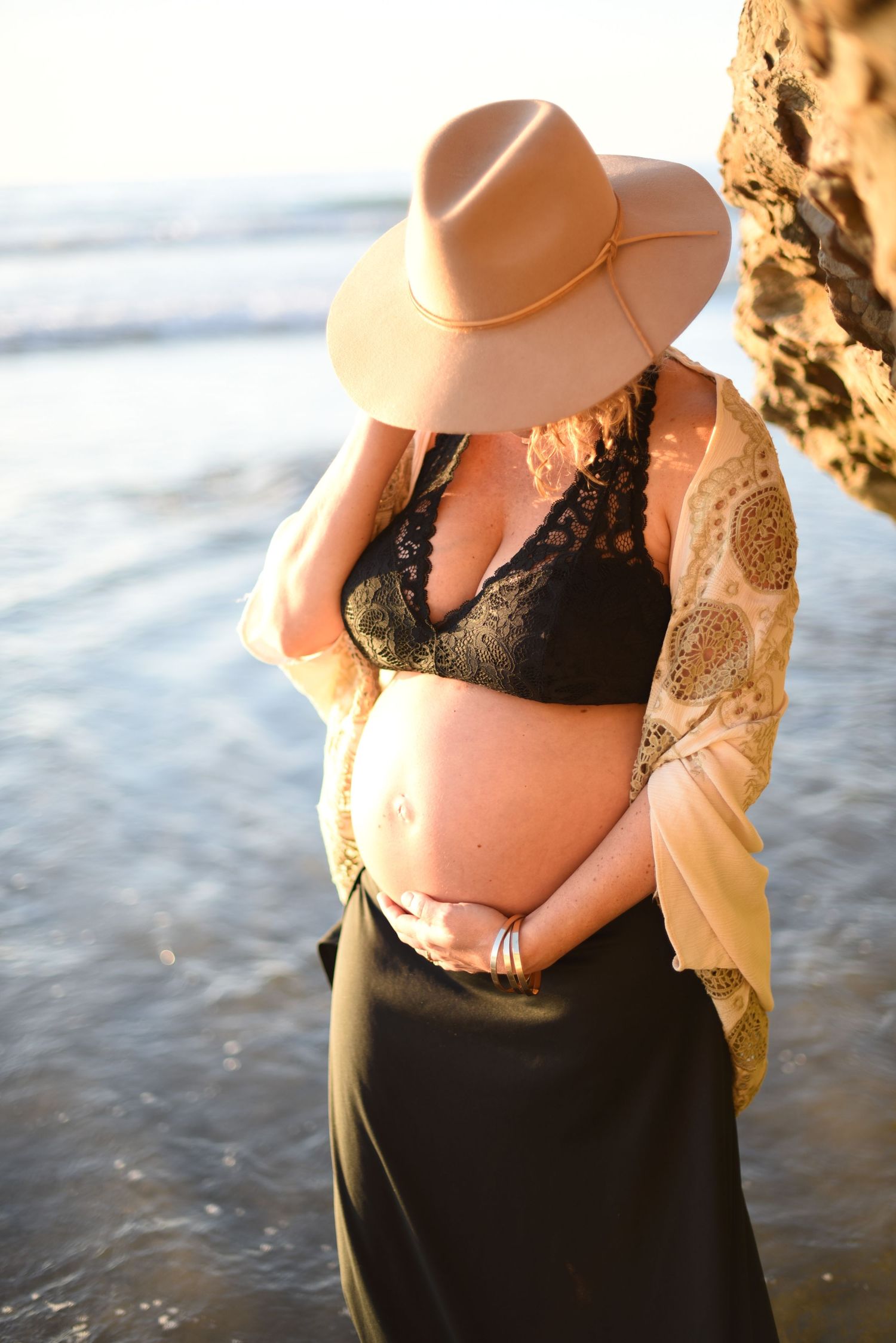 Maternity photoshoot at the beach shows a pregnant woman in a black swimsuit and beige cardigan at sunset.