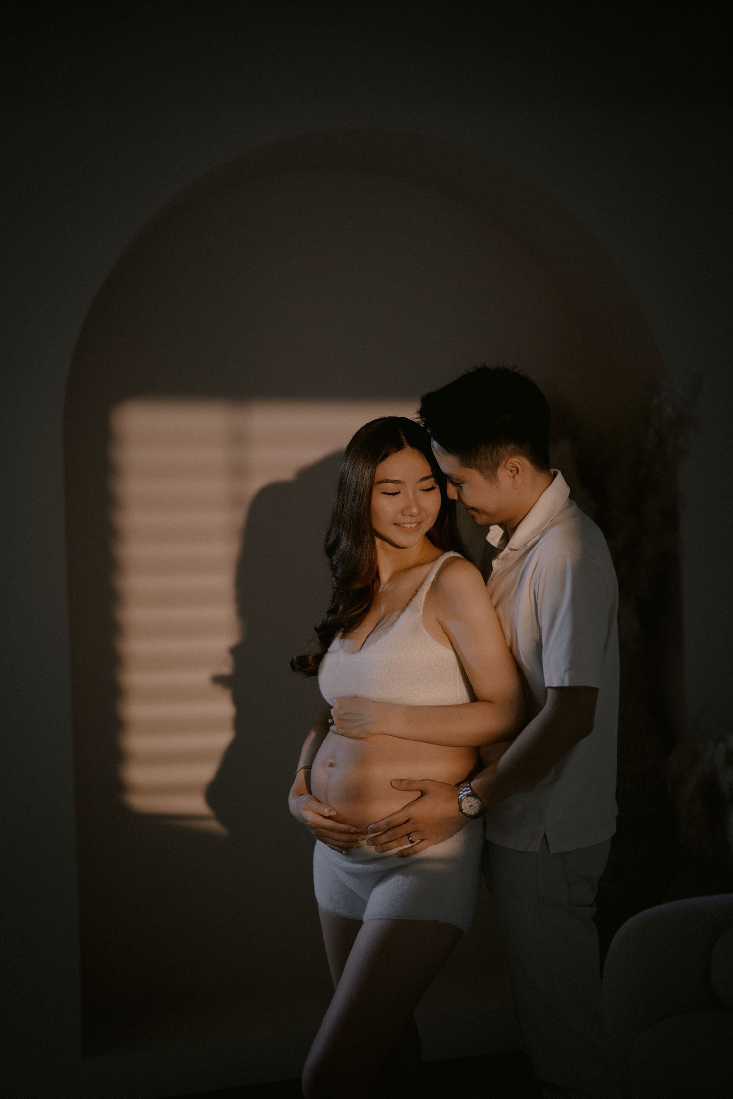 A romantic maternity photoshoot shows a couple embracing in a dimly lit room with window light casting shadows.