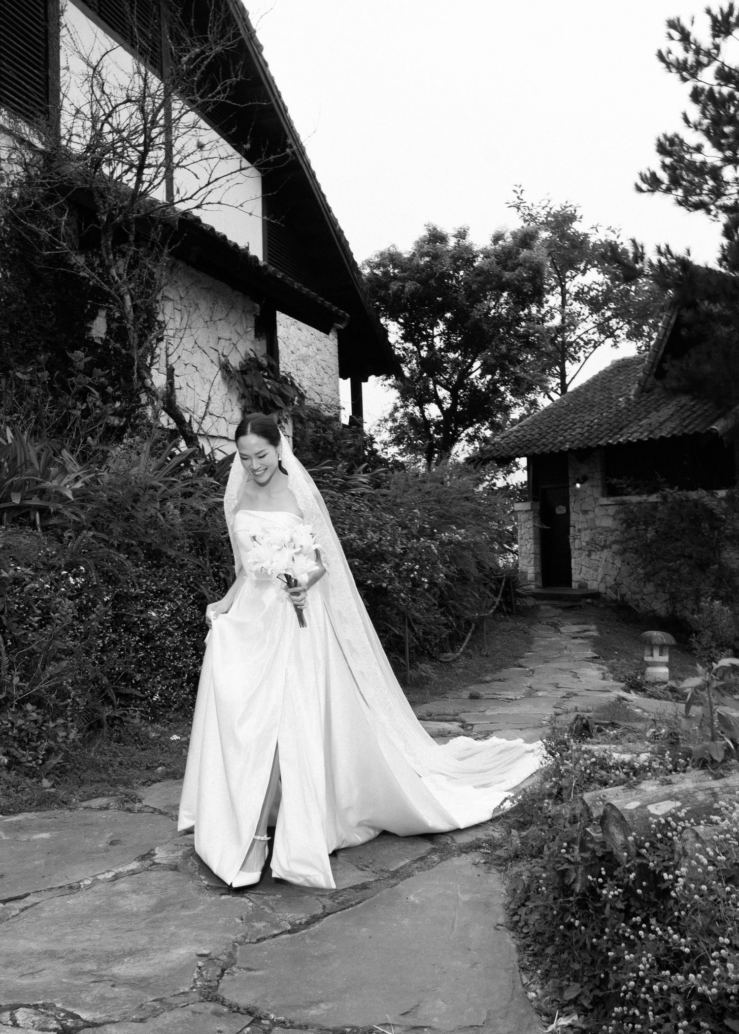 Black and white photo of bride in flowing wedding gown with long veil outdoors.
