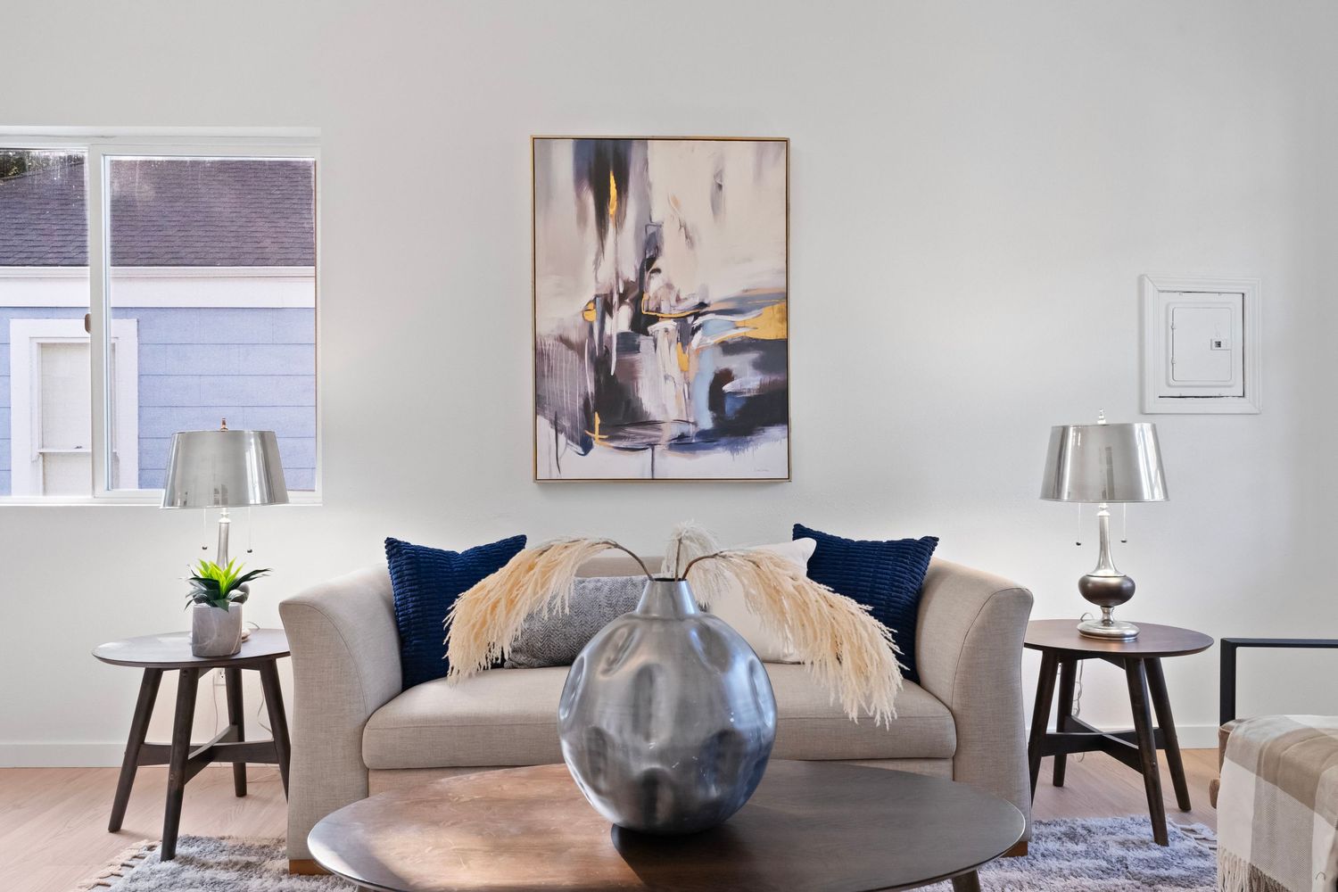 Modern living room staging with navy blue accent pillows, round side tables, and neutral artwork displayed on white walls.