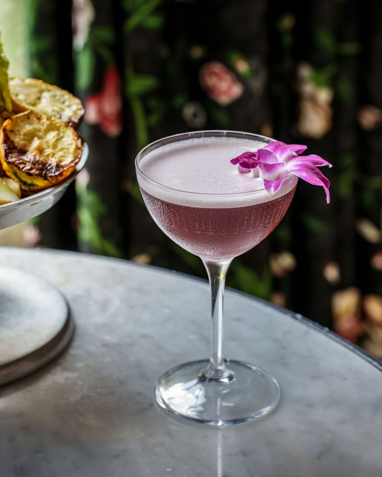 A Commodore Perry purple cocktail garnished with tropical flowers served in a coupe glass on a marble surface in moody lighting.