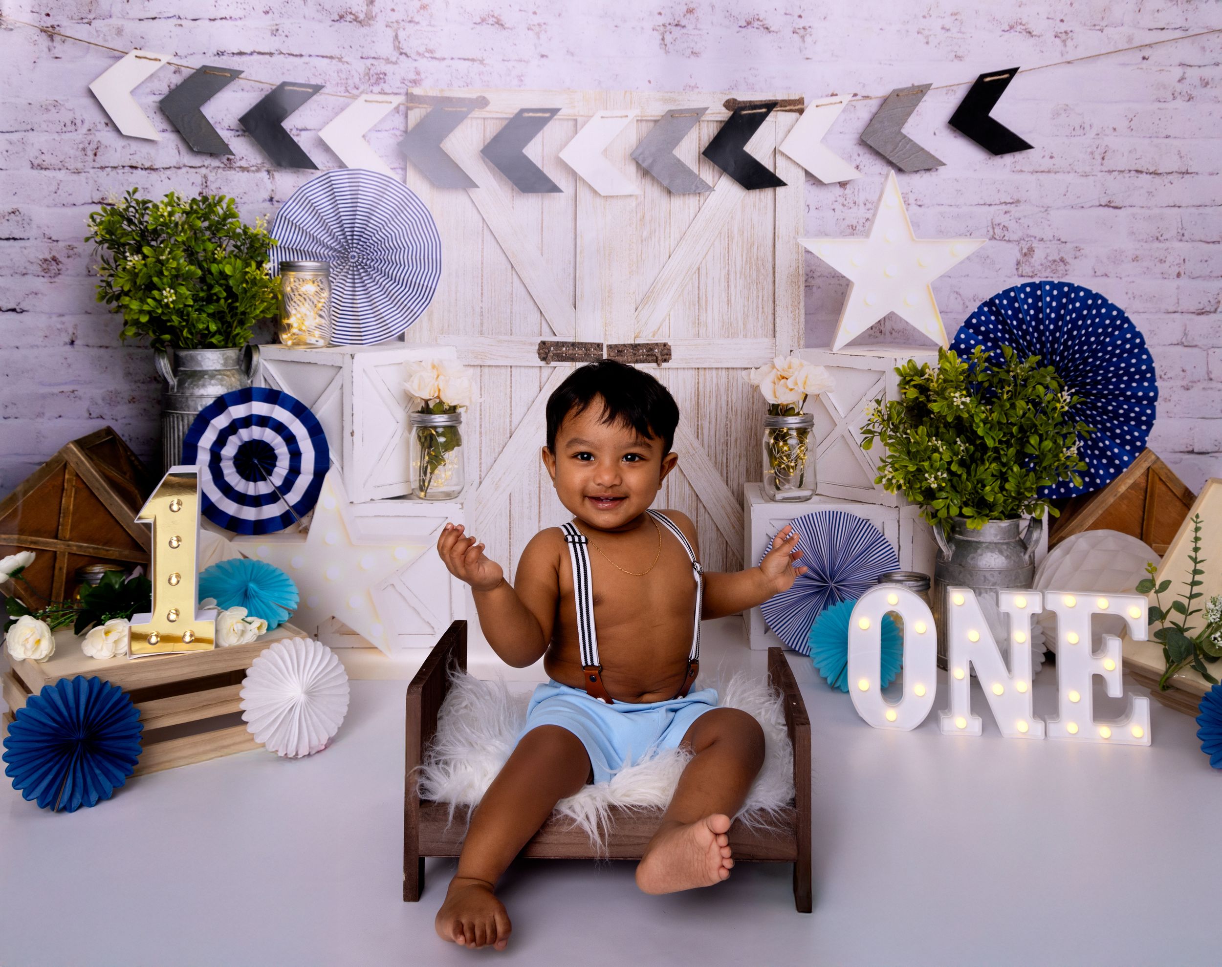 First birthday photo setup with navy and white decorations including paper fans, arrows, and 'ONE' letters against brick wall.