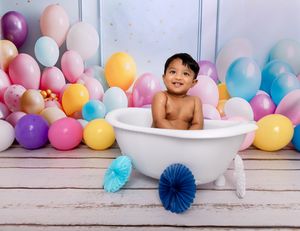 Colorful party balloons and paper fans create a festive backdrop for a playful bathtub photography session.