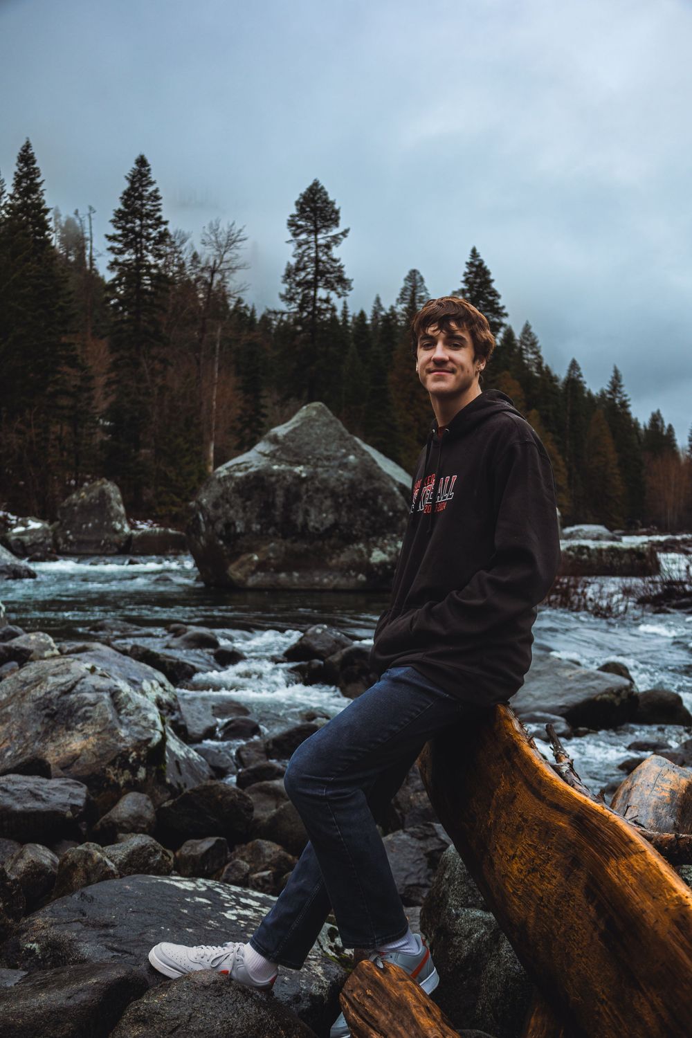 Snow-covered rocks and fallen logs line a rushing river beneath towering evergreen trees.