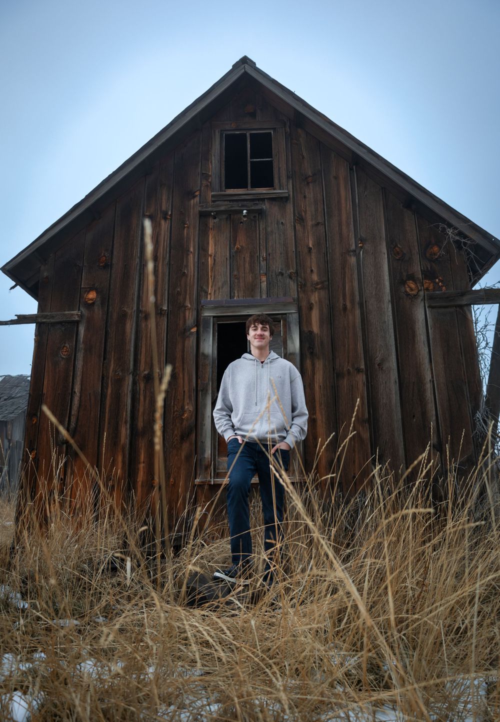 An old weathered wooden barn stands in tall dry grass with snow patches on the ground.