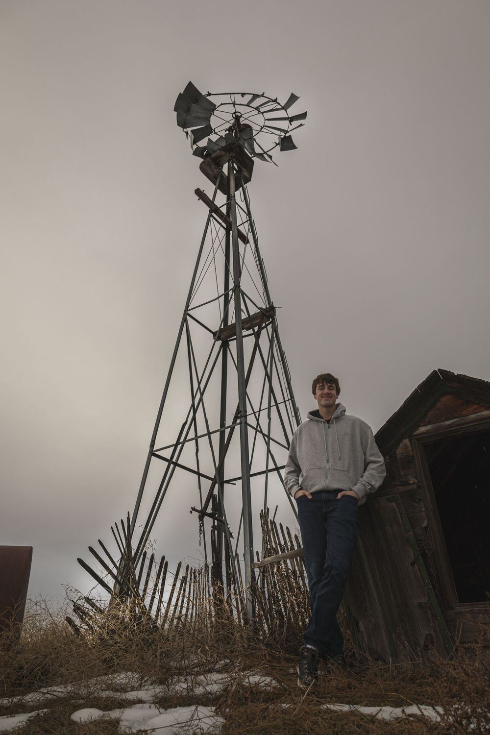 A tall windmill tower stands against an overcast sky with a figure in a light hoodie beside it.