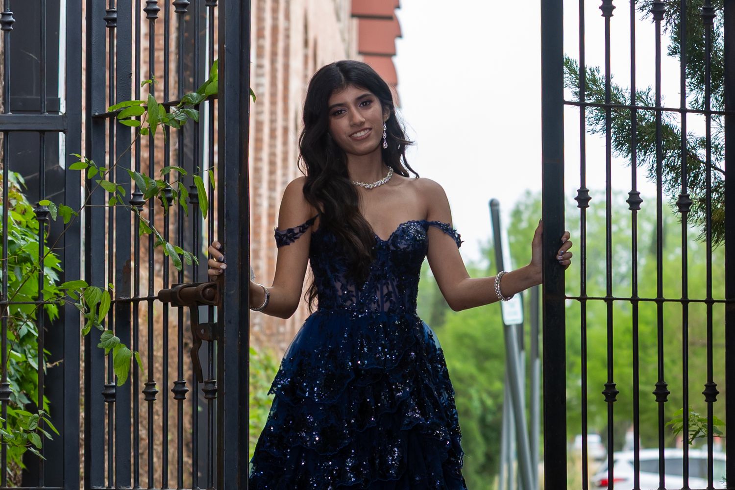 Woman in a sparkly navy blue dress poses elegantly against ornate iron gates in a garden setting.