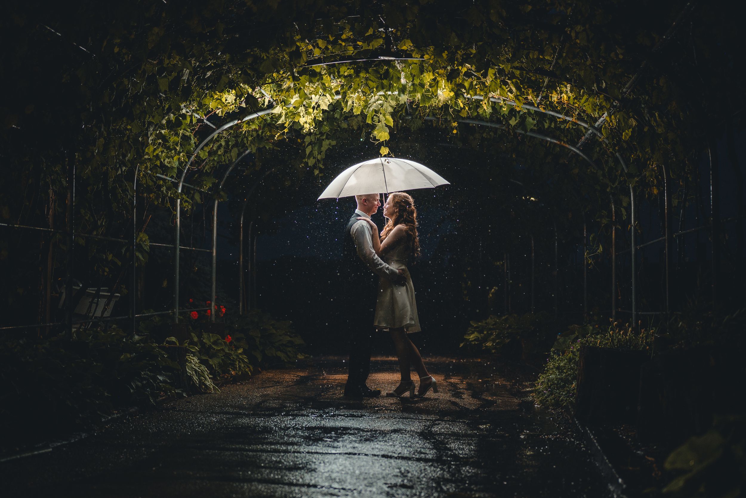 Couple sharing romantic moment under illuminated garden arch with white umbrella at night.