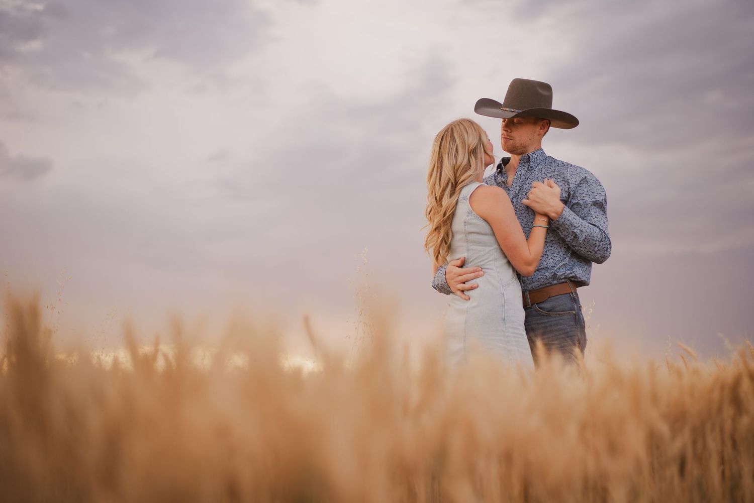 A romantic couple's photoshoot in a wheat field during golden hour with dramatic cloudy skies.