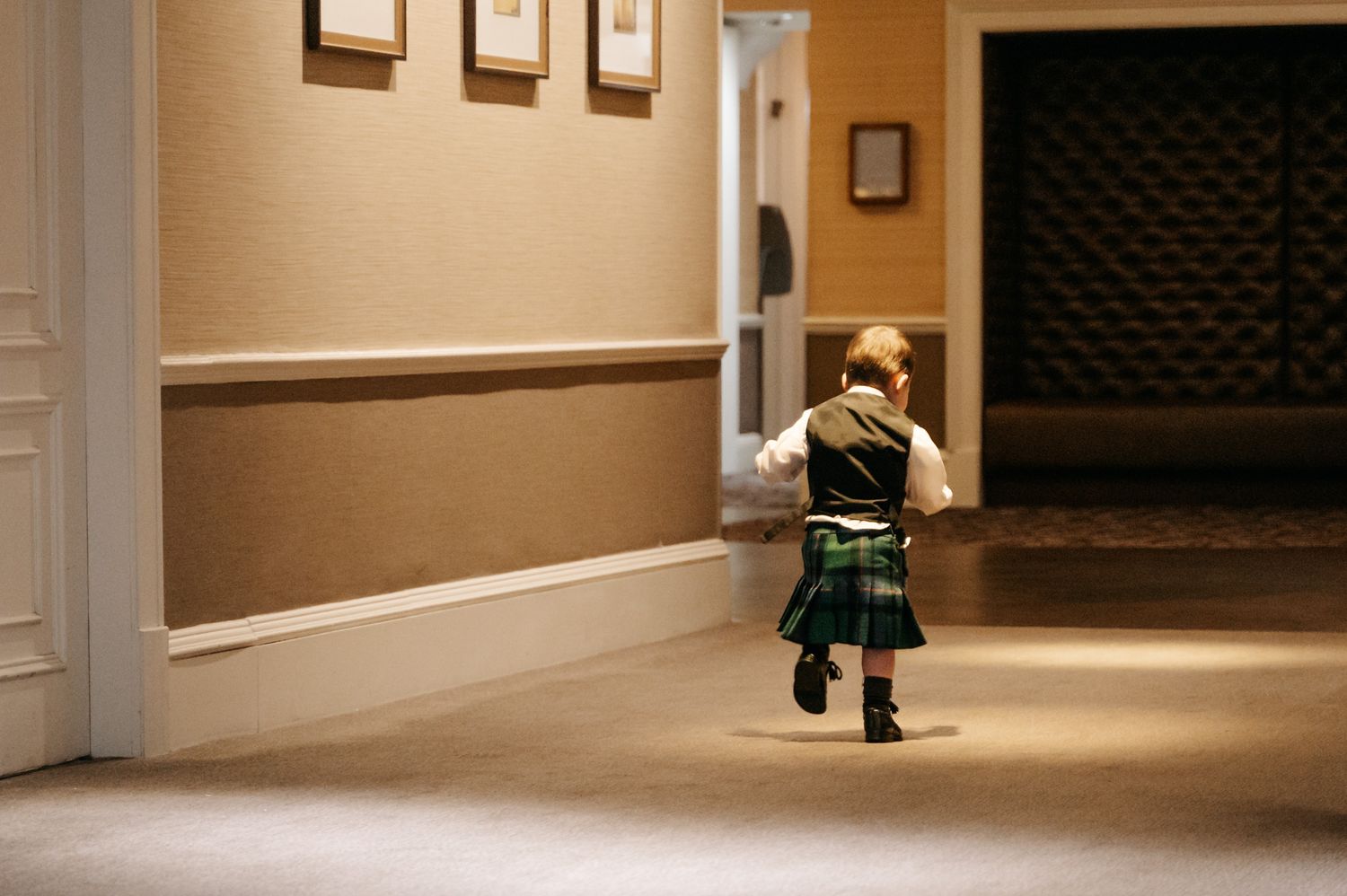 Long hallway with warm lighting and framed artwork on beige walls with small boy running in a kilt.