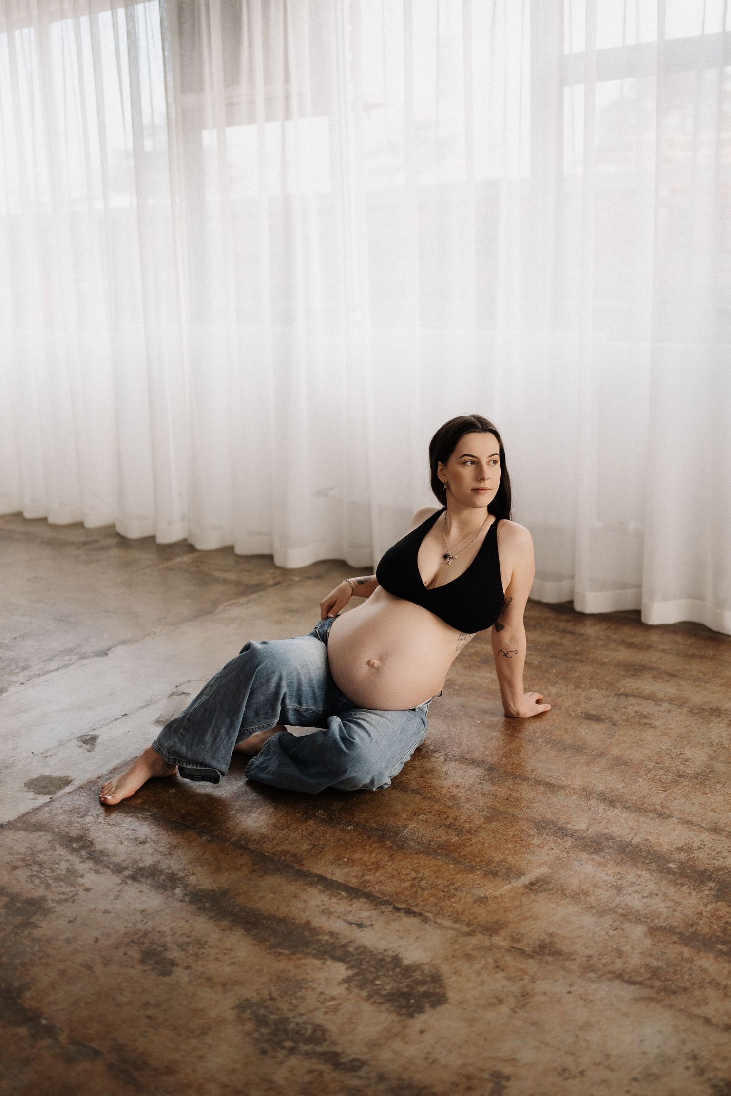 Maternity photography session showing woman in black top and jeans lounging on rustic wooden floor near sheer curtains.