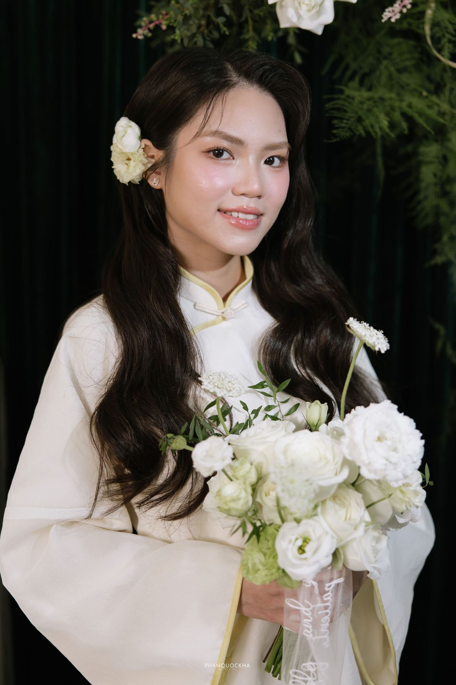 Young Vietnamese woman in white ao dai holding a bouquet of white roses with a flower in her hair.