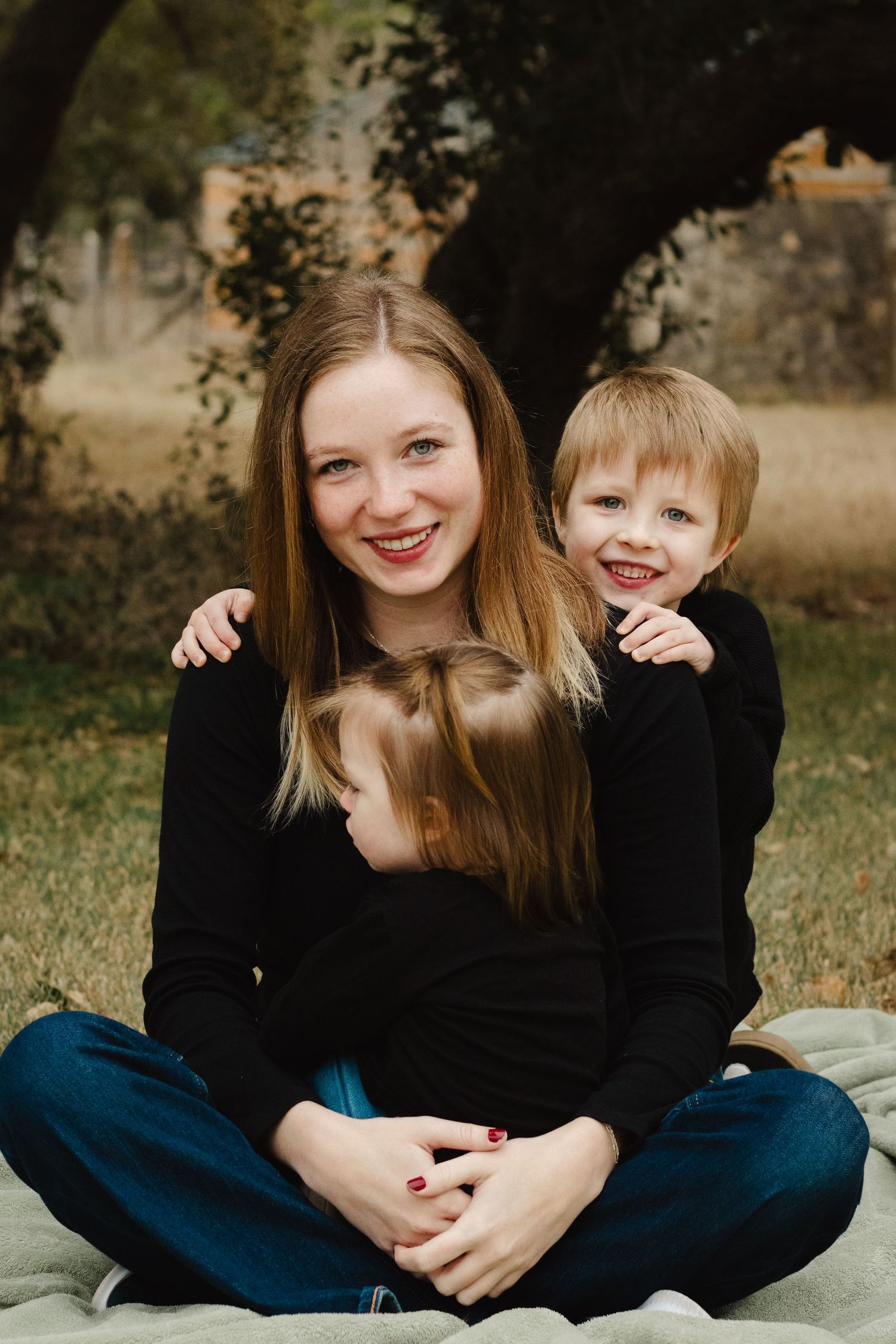 Three people wearing black tops sit together on a blanket outdoors during a casual family photo session.