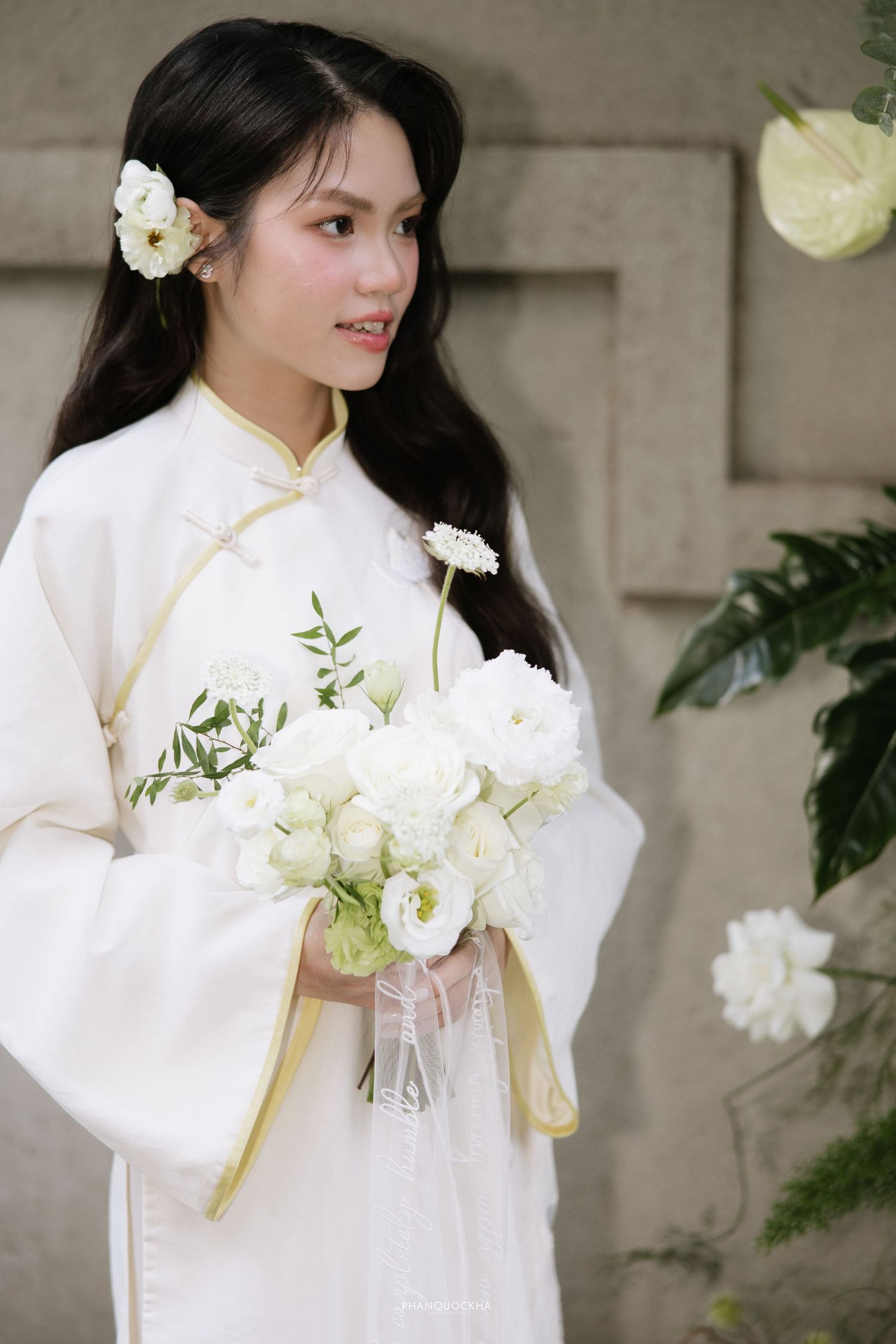 Elegant bride in white Vietnamese ao dai with white flowers in hair holding bouquet.